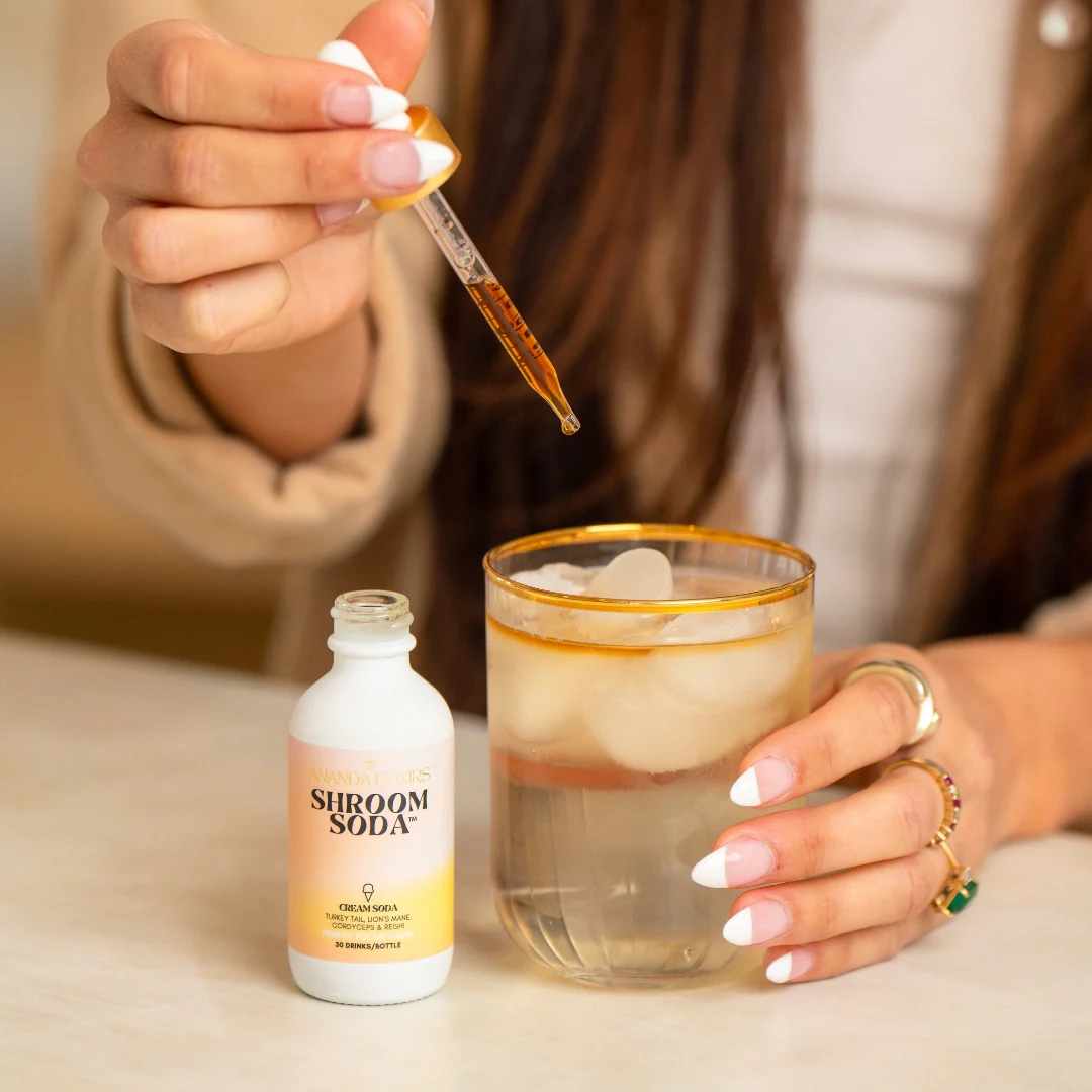Person adding a dropper of Shroom Soda Cream Soda to a glass of Shroom Soda.