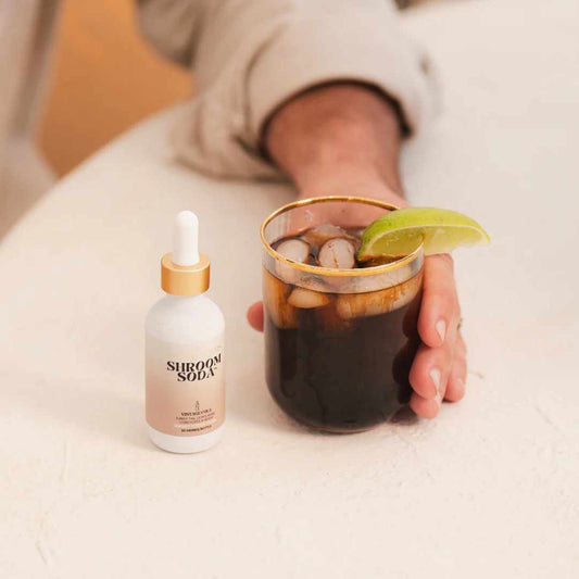Person holding a glass of Vintage Cola Shroom Soda with a lime wedge, next to a bottle labeled 'Shroom Soda'.